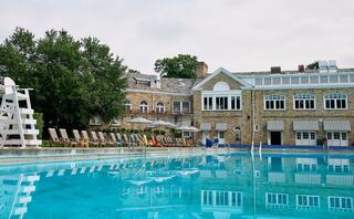 A serene pool area features lounge chairs beside a crystal-clear water, with a charming stone building and trees in the background.
