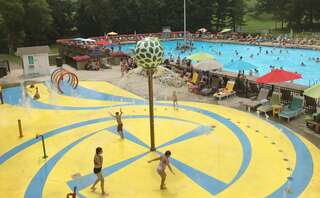 A busy pool area with families enjoying the water, colorful umbrellas, and a playful water feature for children under a sunny sky.