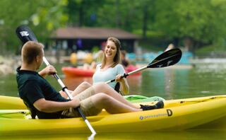 Two people enjoy a fun kayaking moment on a calm lake, smiling and engaging with each other amidst a lush, green backdrop.