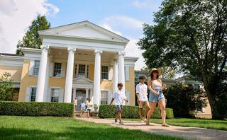 A family strolls on a pathway in front of a grand, yellow mansion with white columns, surrounded by lush greenery and a blue sky.