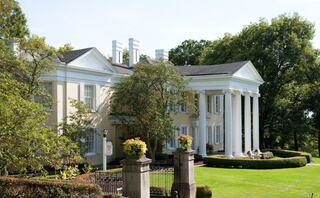 A grand, elegant mansion features white columns, manicured gardens, and lush greenery, exuding classic architectural beauty.