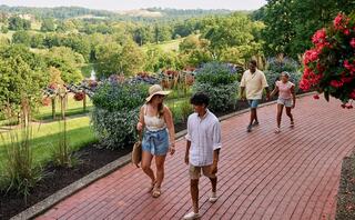 A group of four people strolls along a brick path adorned with vibrant flowers, surrounded by lush greenery and rolling hills.