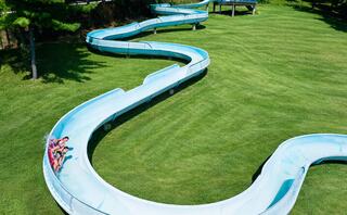 A winding water slide meanders through lush green grass, with two people enjoying a ride down its curves under a clear blue sky.