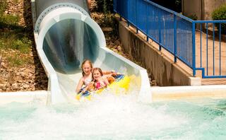 Two children joyfully slide down a water slide in an amusement park, splashing into the pool below on a sunny day.