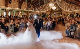 A newlywed couple dances under fairy lights and a chandelier, surrounded by guests in a beautifully decorated reception hall.