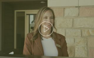 A smiling professional woman in a stylish outfit stands by a stone wall, ready to engage, with a play button in view, indicating video content.