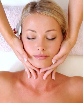 A serene woman enjoys a facial massage, eyes closed, with gentle hands cradling her face and a flower accentuating her hair.