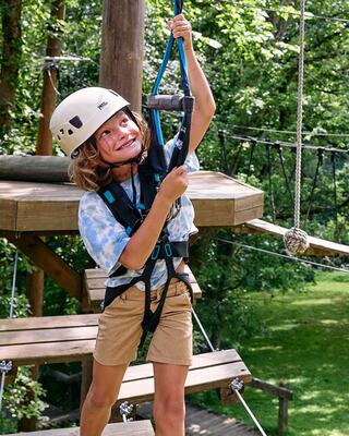 A joyful child in a helmet climbs with a harness on a ropes course, surrounded by lush greenery and wooden structures. Adventure awaits!