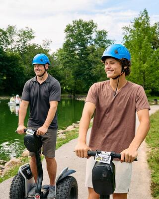 Two young men on segways wear helmets while enjoying a scenic park with a pond. They appear relaxed and cheerful in a sunny setting.