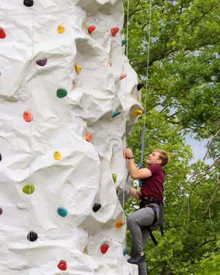 A person climbs a white rock wall, wearing climbing gear, surrounded by lush green trees under clear skies.
