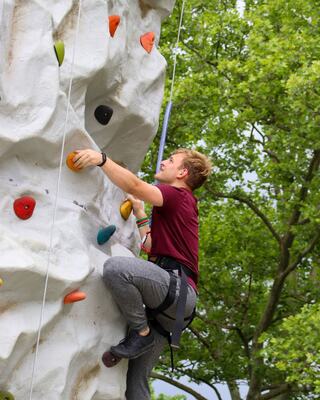 A climber ascends a rock wall, focusing intently while harnessed and surrounded by lush green trees under a cloudy sky.