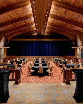 A spacious hall features round tables with black tablecloths, wooden accents, and a stage draped in blue curtains, ideal for events.