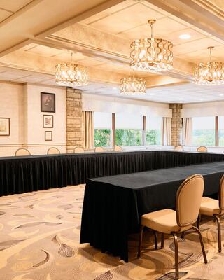 A spacious meeting room features elegant chandeliers, long black tables, and soft lighting, with windows offering a view of greenery outside.