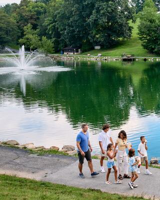 A family walks along a path by a serene pond, enjoying each other's company amidst lush greenery and a fountain in the background.