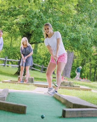A group of friends enjoys mini-golf outdoors, with one woman preparing to take a shot while others watch and relax in a lush setting.