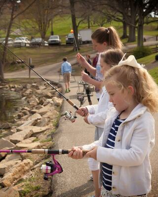 A group of children and adults fish by a pond, enjoying a sunny day together. The atmosphere is joyful and relaxed, surrounded by nature.
