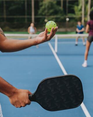 A person holds a bright green ball with one hand and a paddle in the other, ready to play on a blue court with others in the background.