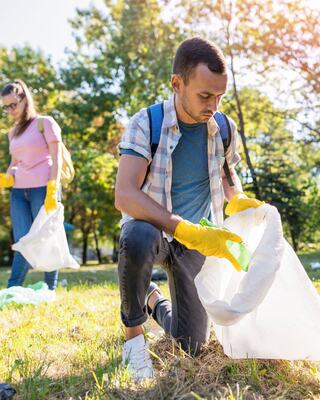 Two individuals are cleaning a park, collecting trash while wearing gloves. The sun shines, highlighting their efforts for a cleaner environment.