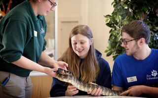 A woman holds a snake while two smiling volunteers observe, showcasing an engaging educational moment about reptiles.