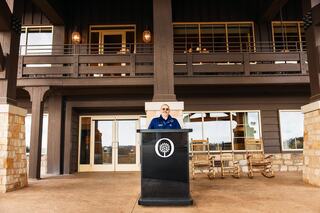 A person stands at a podium in front of a stylish, modern building with large windows and a stone exterior, ready to address an audience.
