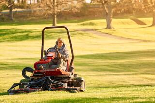 A man operates a riding lawn mower on a sunny day, expertly cutting grass in a lush green landscape. Trees line the background.