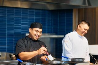 A smiling chef in a black uniform uses a spatula, while another chef in white looks on, both in a modern kitchen.