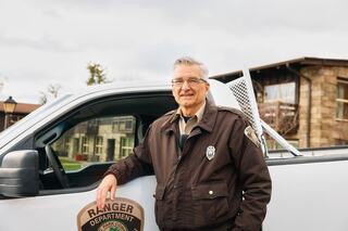 A senior park ranger stands proudly beside his truck, wearing a uniform, showcasing a badge, with a charming, welcoming expression.