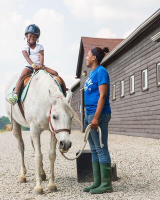 A young girl rides a white horse, smiling happily, while an adult companion stands beside her, holding the horse's lead.