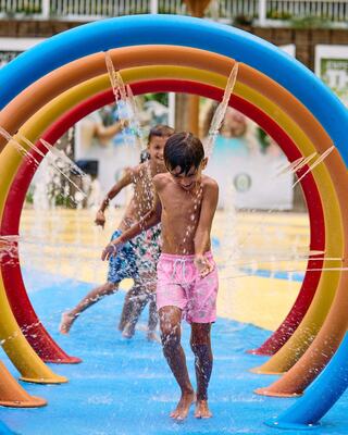 Children joyfully run through colorful water arches, splashing and playing in a vibrant splash pad on a sunny day.