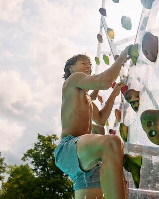A shirtless young man climbs a colorful rock wall, smiling with determination, against a backdrop of trees and a bright sky.