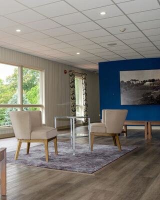 A cozy, modern space features two armchairs, a small table, and large windows with natural light, accented by a blue wall and artwork.