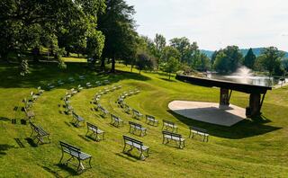 An outdoor amphitheater features a circular arrangement of benches on manicured grass, with a gazebo and a fountain in the background.