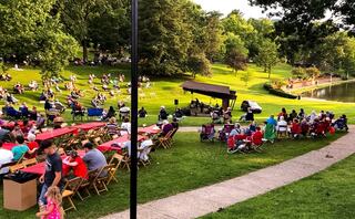A lively outdoor concert in a park with a crowd seated on chairs, enjoying music and food, surrounded by greenery and a serene pond.