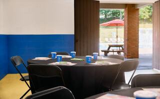 A circular black table is set with plates and cups, viewed from a room leading to a pool area with an umbrella and chairs outside.