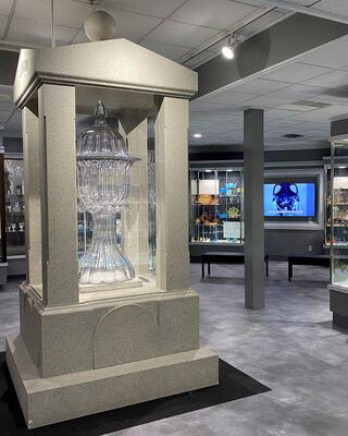 A large, ornate glass sculpture is displayed inside a modern exhibit space, featuring sleek walls and various showcase cases.