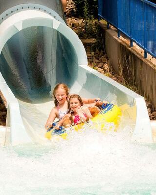 Two joyful girls ride a yellow tube down a water slide, splashing into the pool below while enjoying a sunny day at the water park.