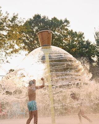 Children play under a large, flowing water feature resembling an ice cream cone, splashing joyfully in the sunlit, aquatic scene.