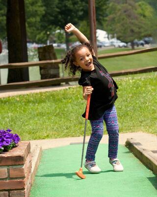 A joyful girl raises her arm in celebration while playing mini-golf, dressed in a black shirt and colorful leggings. Bright and cheerful day.