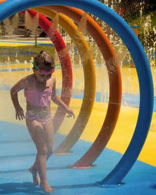 A young girl plays joyfully under colorful water arcs at a bright water park, enjoying a refreshing summer day.