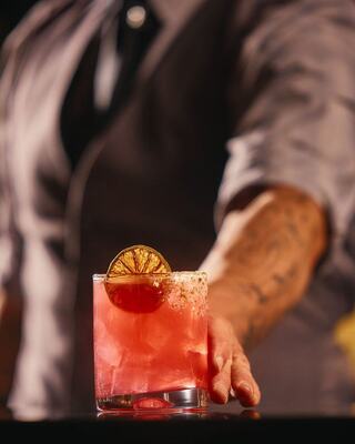 A bartender presents a vibrant cocktail garnished with a dried orange slice, set against a softly lit background.
