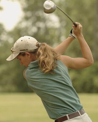 A woman with long hair swings a golf club, dressed in a sleeveless top and cap, focused on her shot in an outdoor setting.