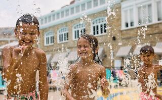 Three joyful children play in water, splashing and laughing under the sun at a poolside, surrounded by a cheerful summer atmosphere.