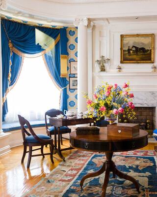 A bright, elegant room features blue drapes, a round table with flowers, and classic furnishings, showcasing a cozy, inviting atmosphere.