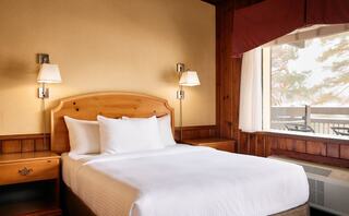 A cozy bedroom features a wooden bed with white linens, bedside lamps, and a large window with a view, creating a warm, inviting atmosphere.