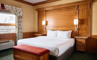 A cozy bedroom features a queen bed with white linens, wooden accents, bedside lamps, a plush red bench, and a window with patterned curtains.