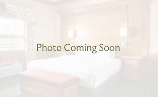 A cozy bedroom features a large bed with white linens, wooden accents, bedside lamps, and a window with patterned curtains.