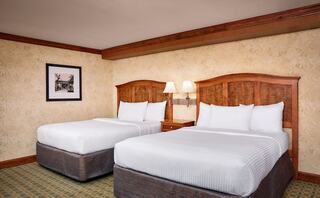 A cozy hotel room features two neatly made beds, wooden accents, a simple lamp, and a framed picture on the wall.