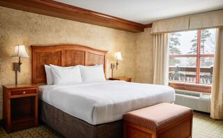 A cozy hotel room features a queen bed with white linens, wooden furnishings, and a large window with views of nature outside.