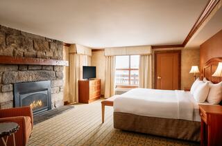 Cozy bedroom featuring a plush bed, a stone fireplace, a TV, and large windows with natural light, enhancing a warm, inviting atmosphere.