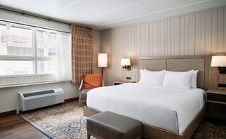 A cozy, modern hotel room features a large bed, soft lighting, an accent chair, and bright curtains, with warm wooden tones and a rug.